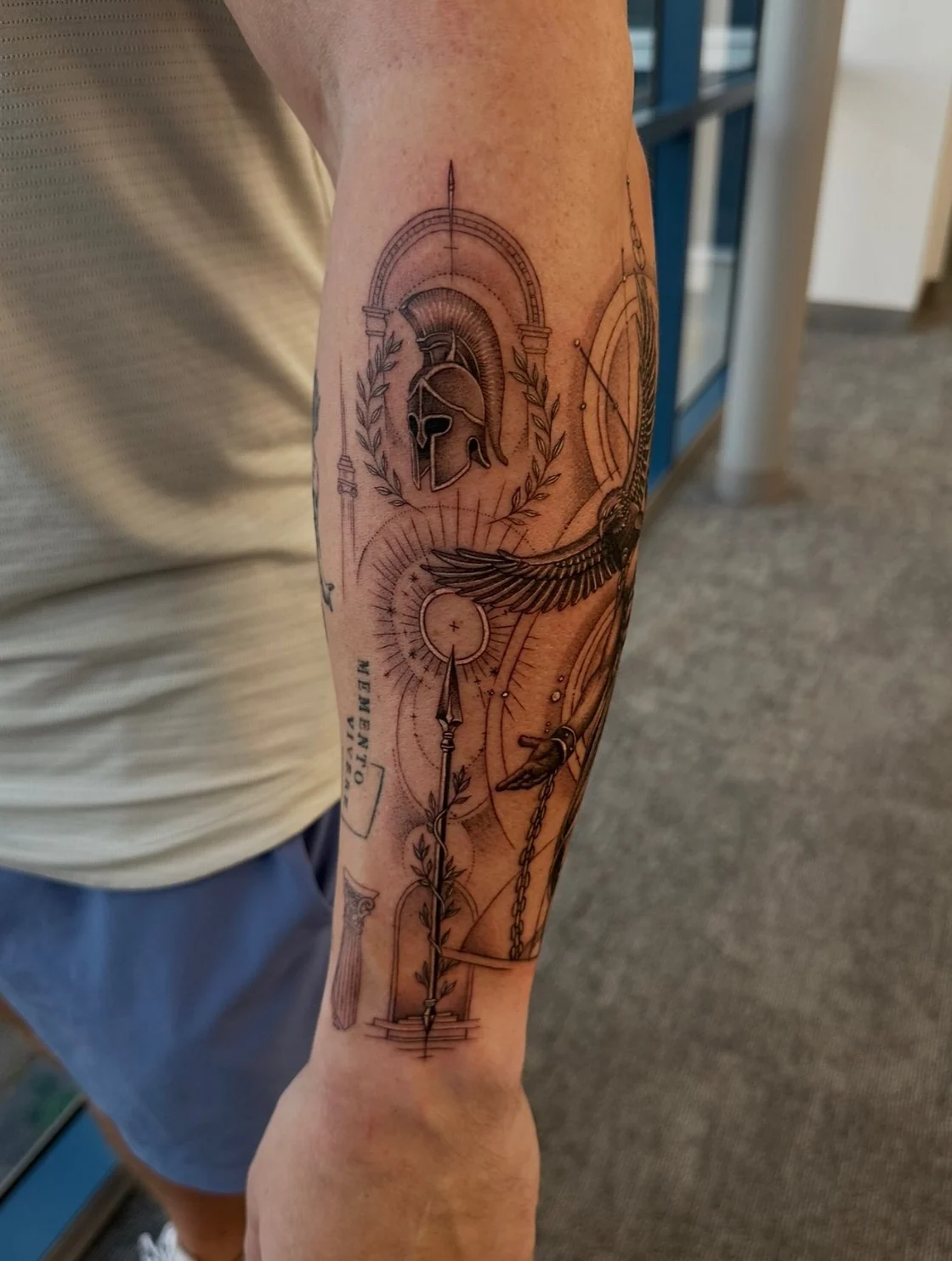 A shaded Spartan helmet anchors a forearm composition with a laurel wreath, circular details, and crisp geometric structure. Tattoo work by Brian of Vicious Ink.