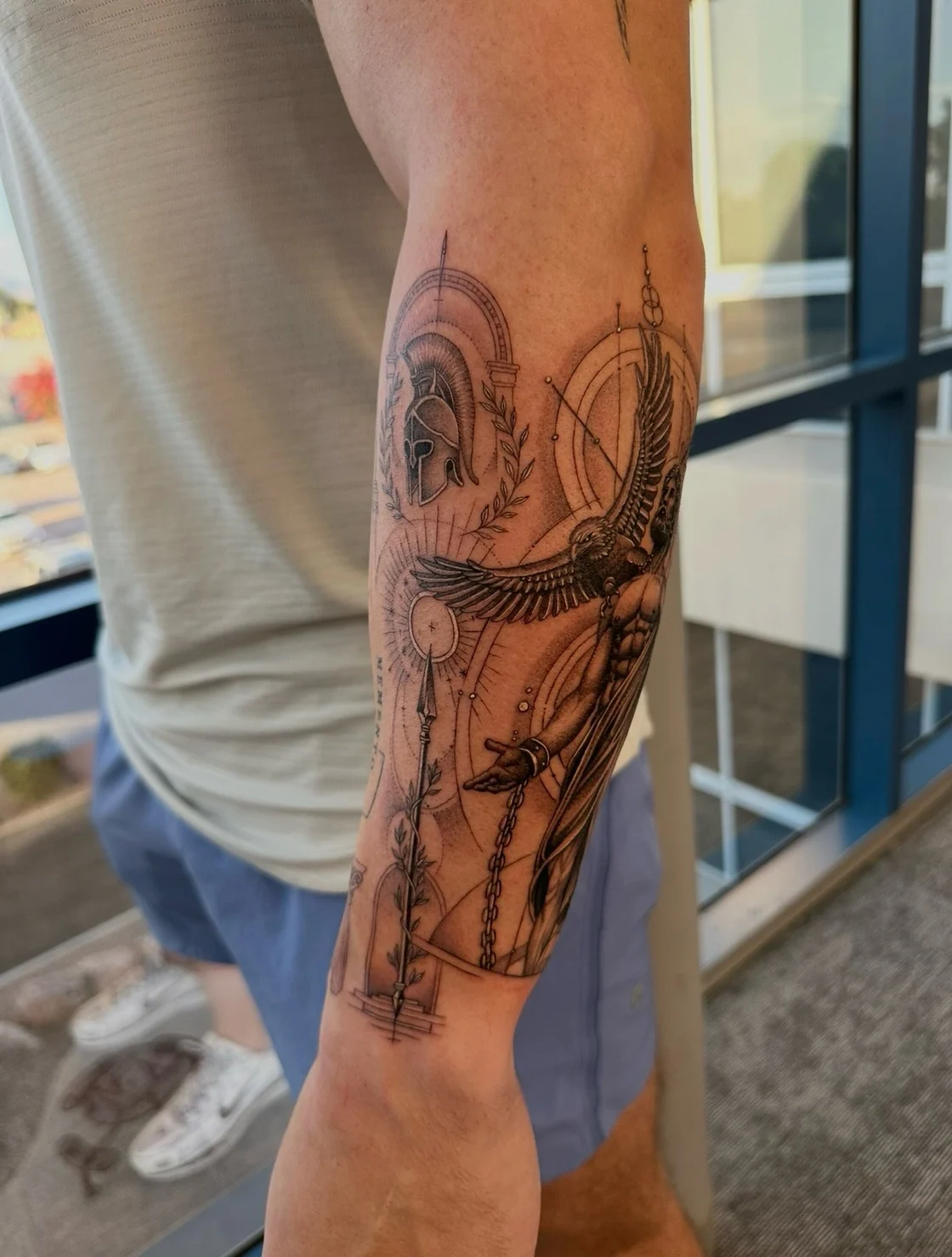 A black and grey forearm composition combines a classical helmet, outstretched eagle wings, a sword, laurel leaves, and fine geometric circles. Tattoo work by Brian of Vicious Ink.