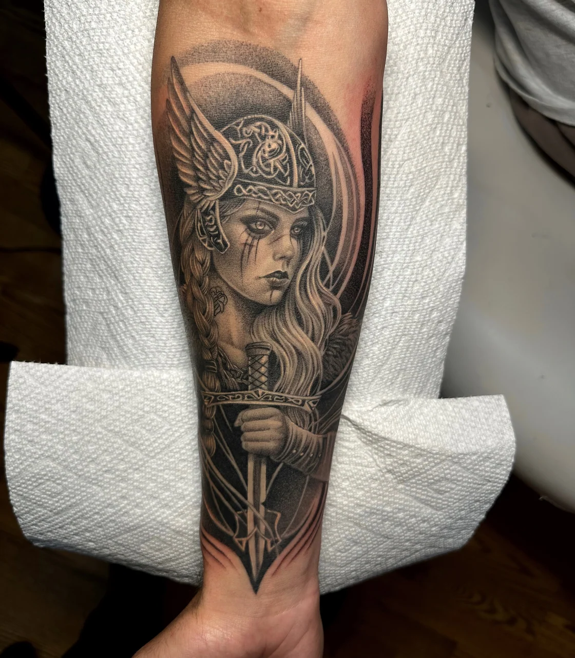 A black and grey female warrior portrait combines a helmet, long hair, and sweeping ornamental curves on the forearm. Tattoo work by Brian of Vicious Ink.