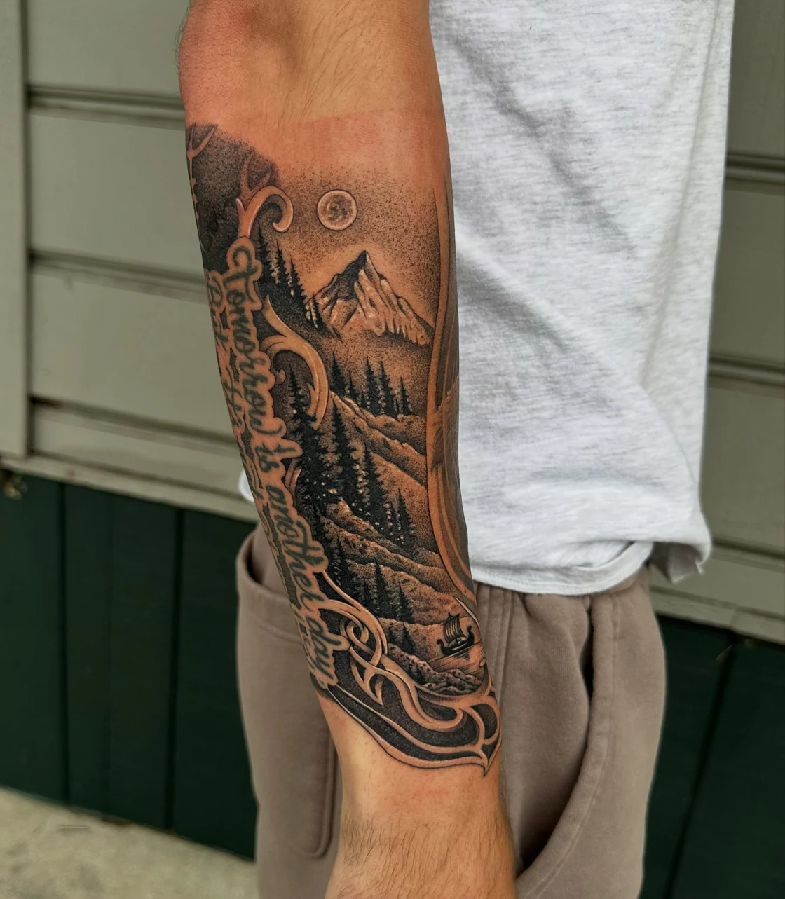 A black and grey landscape combines layered mountains, evergreen trees, flowing ornamental framing, and vertical lettering. Tattoo work by Brian of Vicious Ink.