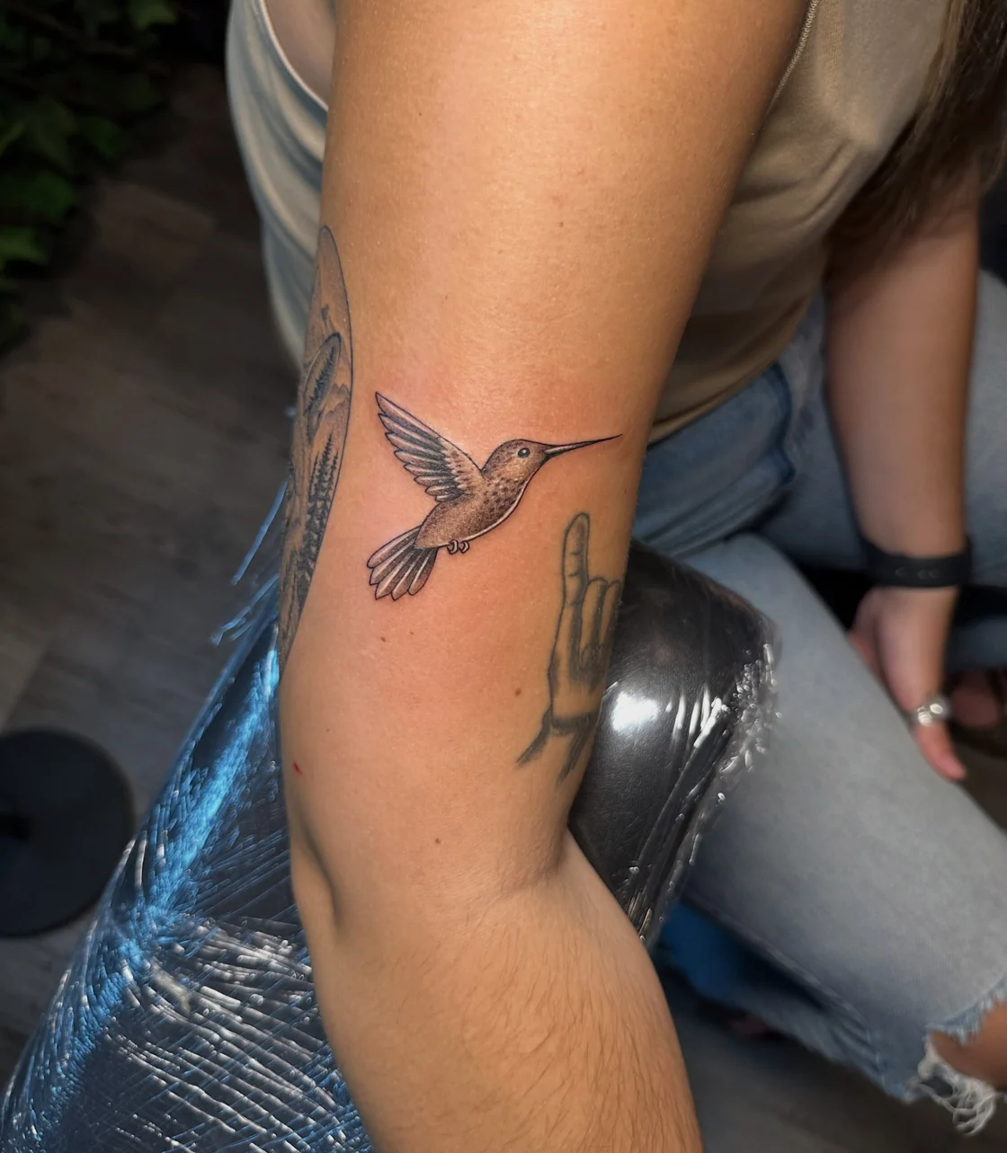 A small hummingbird tattoo on the arm uses delicate wing outlines, fine feather details, and soft grey shading. Tattoo work by Brian of Vicious Ink.