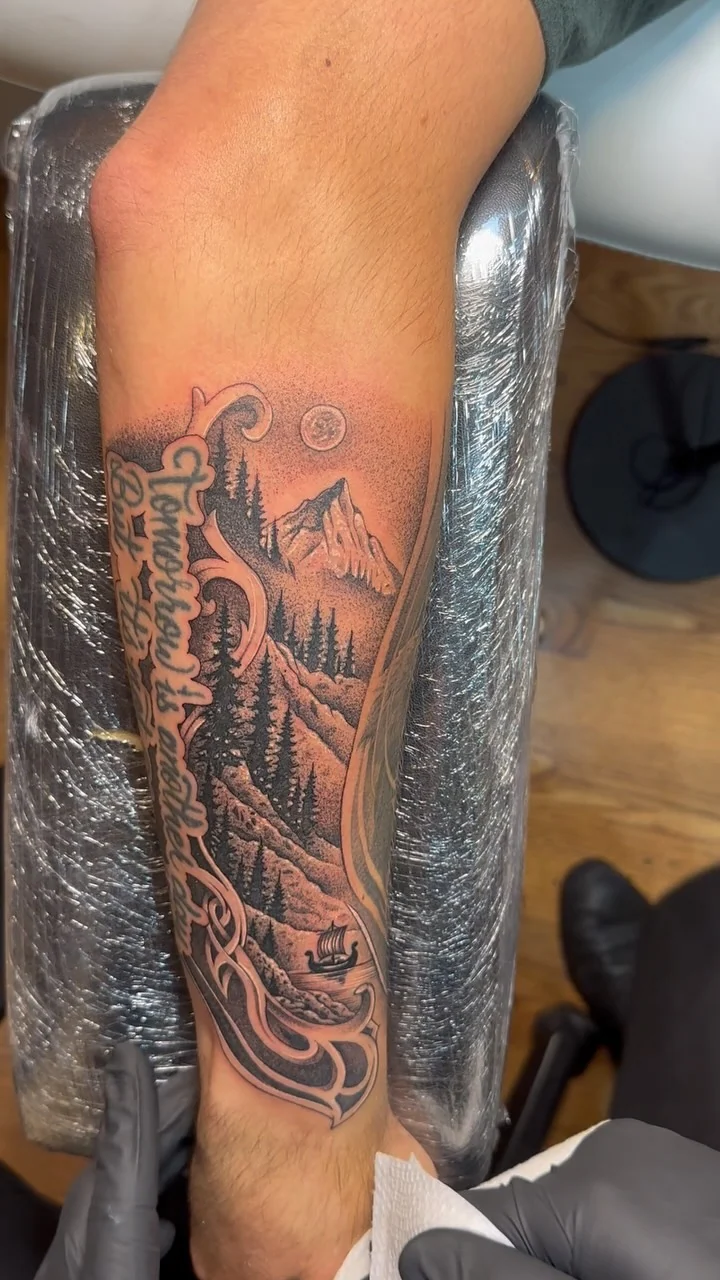 The video poster shows a black and grey mountain landscape with trees and a stream inside an ornate border; the visible side lettering is credited separately. Tattoo work by Brian of Vicious Ink.