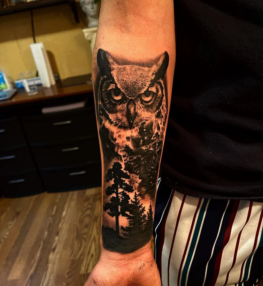 A black and grey owl sits above a forest scene on the forearm, with layered feathers, trees, and strong tonal contrast. Tattoo work by Brian of Vicious Ink.