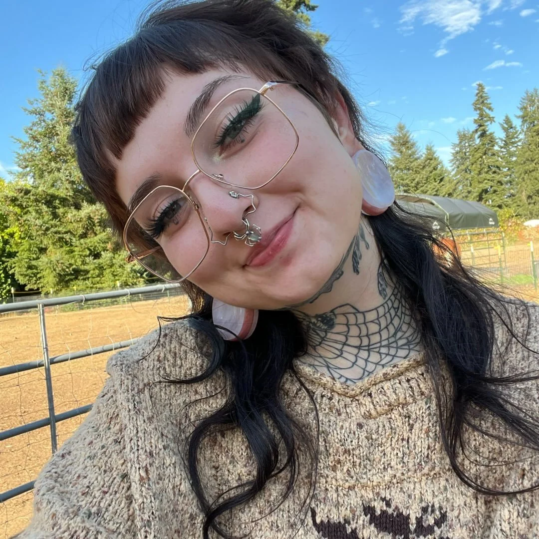 Abra outdoors, wearing glasses and a knit sweater, with evergreen trees and a blue sky behind her.