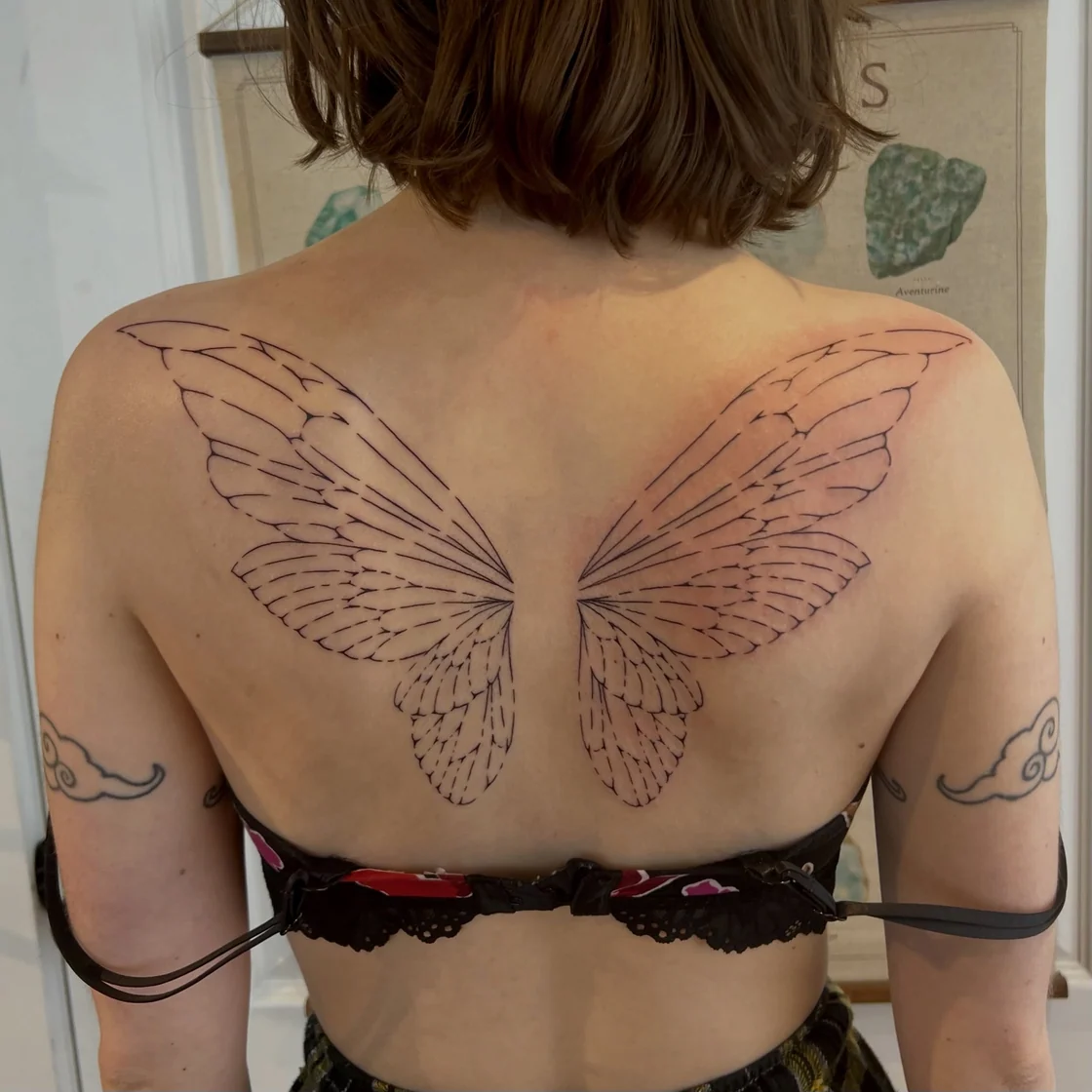 A large back outline tattoo traces symmetrical fairy or butterfly wings across the shoulder blades with open skin and fine line detail. Tattoo work by Abra of Vicious Ink.