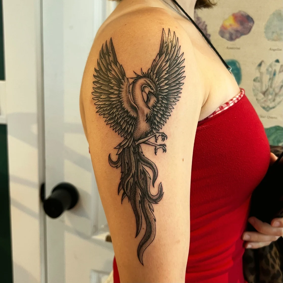 A black and grey phoenix spreads across the shoulder and upper arm with layered feathers, talons, and a long ornamental tail. Tattoo work by Abra of Vicious Ink.