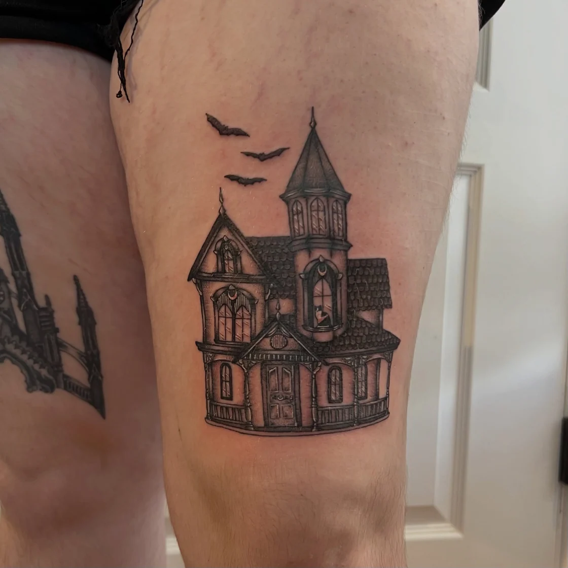 A black and grey haunted house or old building scene sits on the thigh with architectural lines and atmospheric shading. Tattoo work by Abra of Vicious Ink.