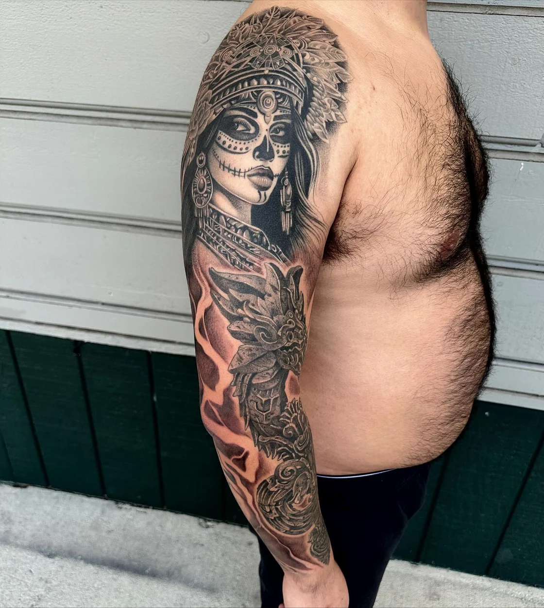 A large black and grey upper arm sleeve combines an Aztec goddess, Quetzalcoatl, feathered forms, and ornate animal details. Tattoo work by Brian of Vicious Ink.